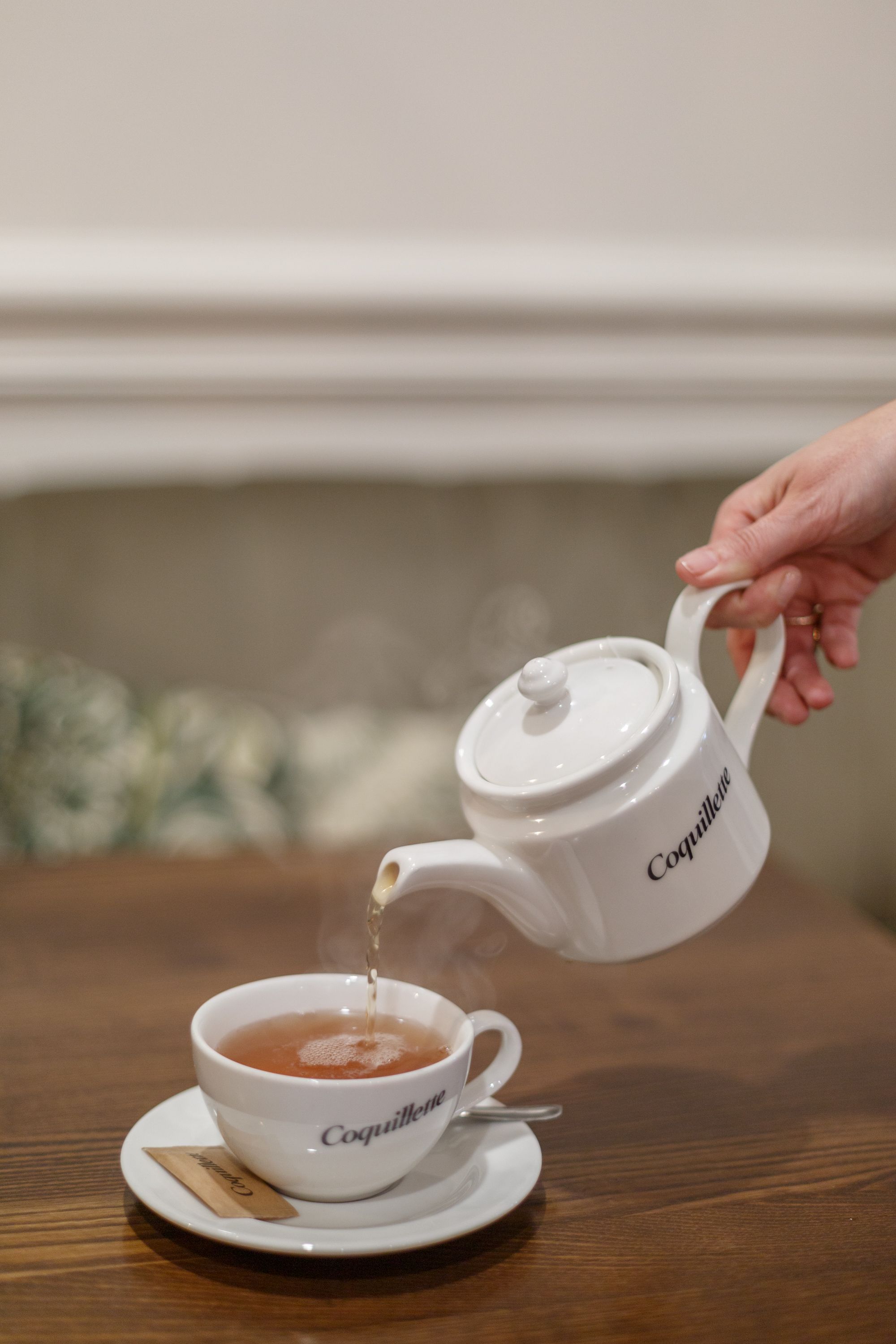 Une main verse du thé fumant d'une théière blanche marquée Coquillette dans une tasse assortie, sur une table en bois devant la cheminée